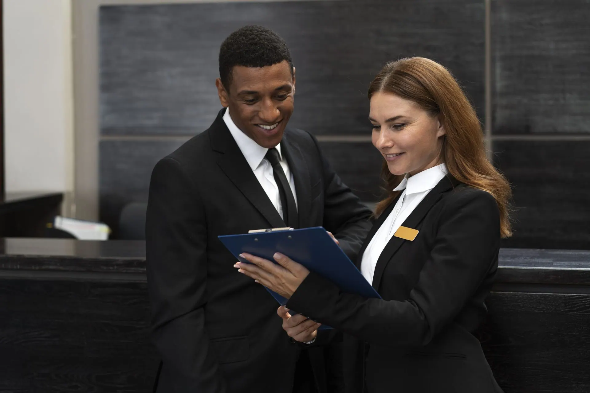 Two front desk staff in dark suits reviewing a clipboard together